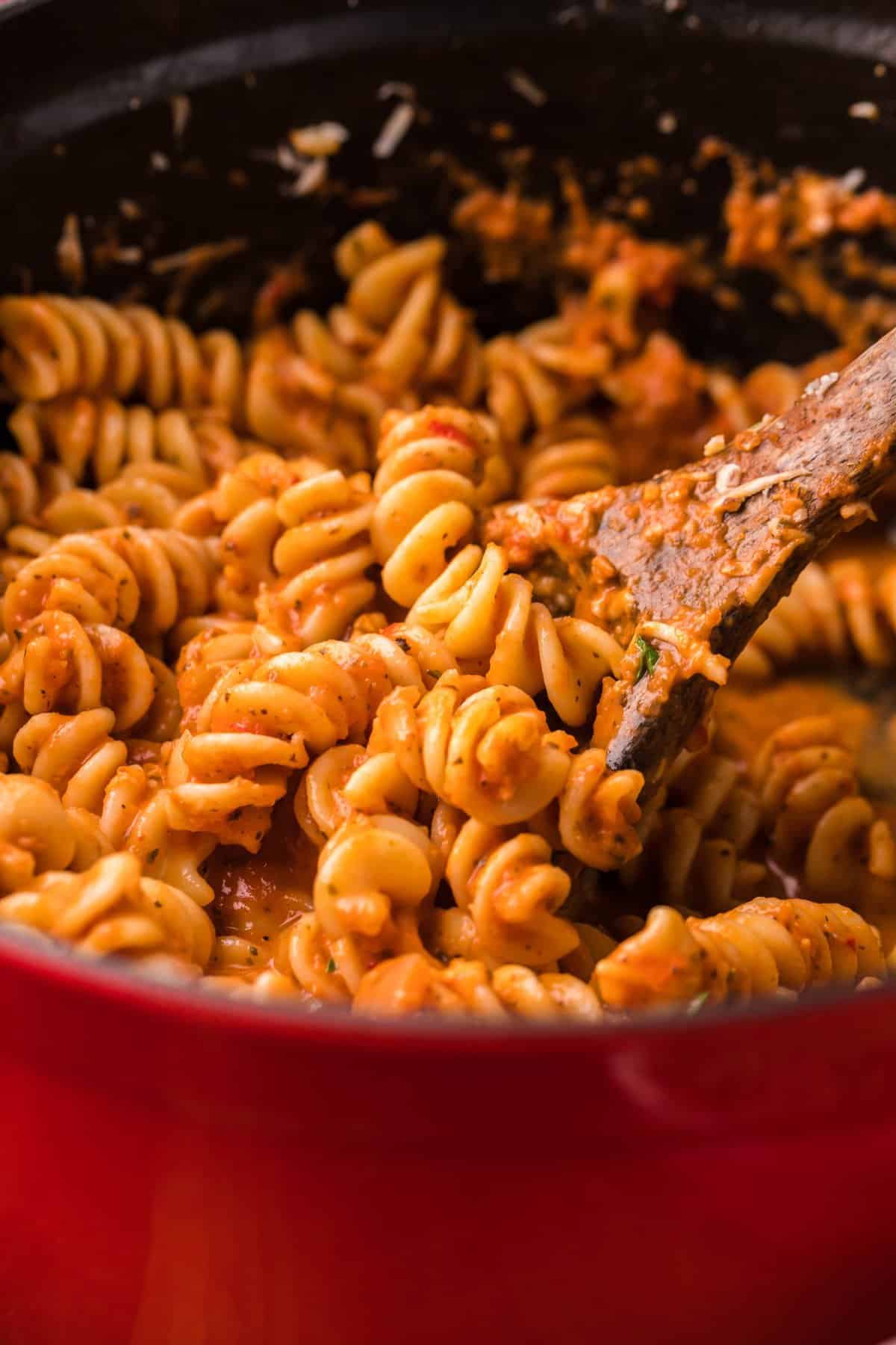 Red pot with spicy fusilli pasta and a wooden spoon taking a portion.