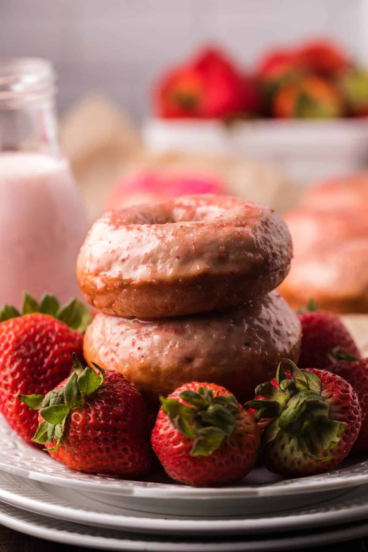Two donuts on white plates surrounded by fresh strawberries.