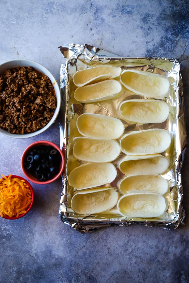 Foil lined tray of taco boats with a bowl of meat, olives, and shredded cheese. 