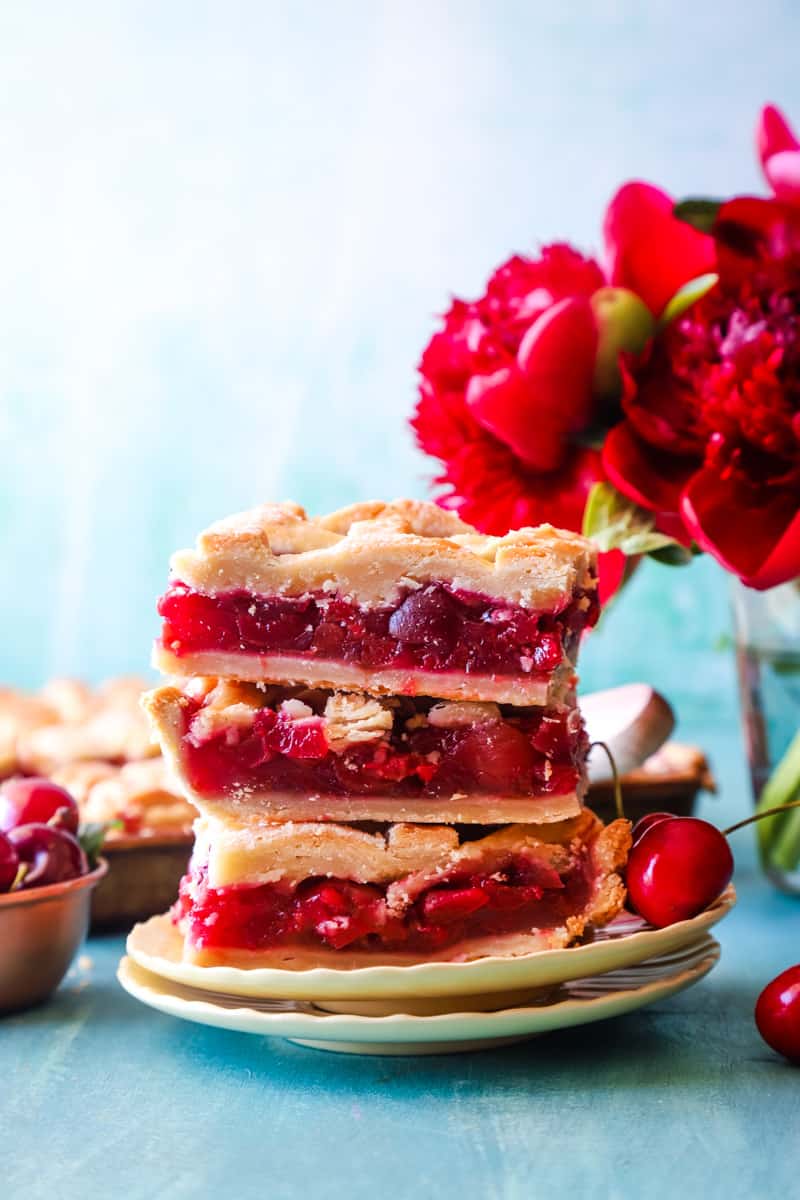 Three cherry slab pie squares stacked on a plate with flowers in the background. 