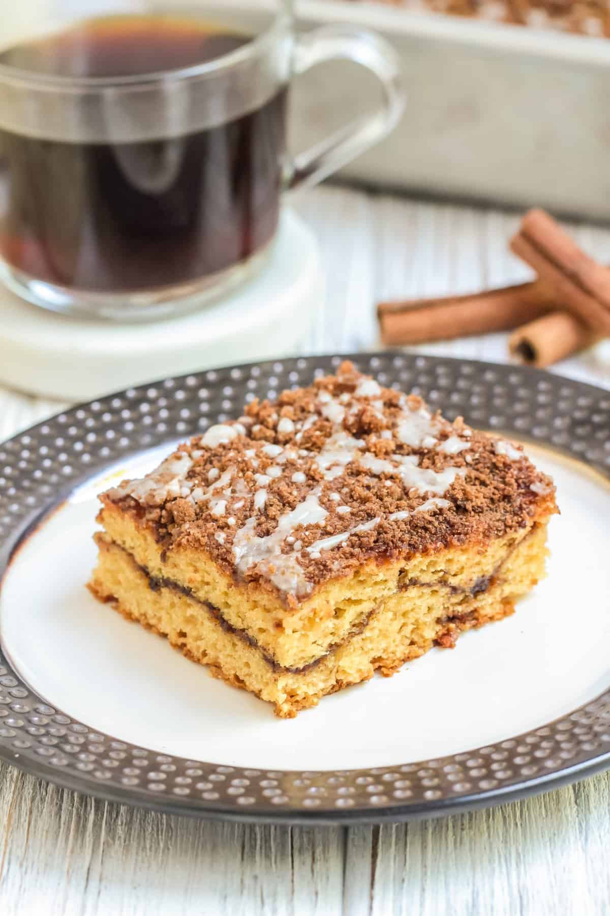 Slice of coffee cake on a white plate with cup of coffee in the background. 