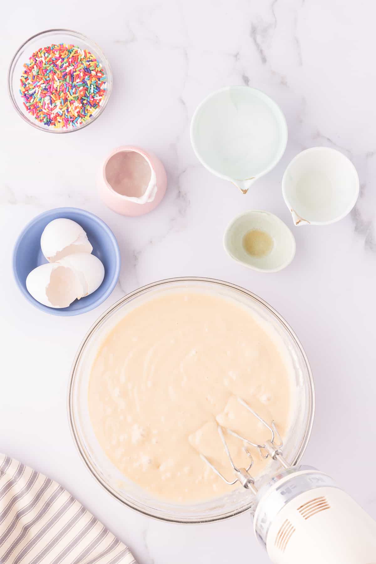 Large glass mixing bowl with vanilla sheet cake batter and bowl of sprinkles next to it. 