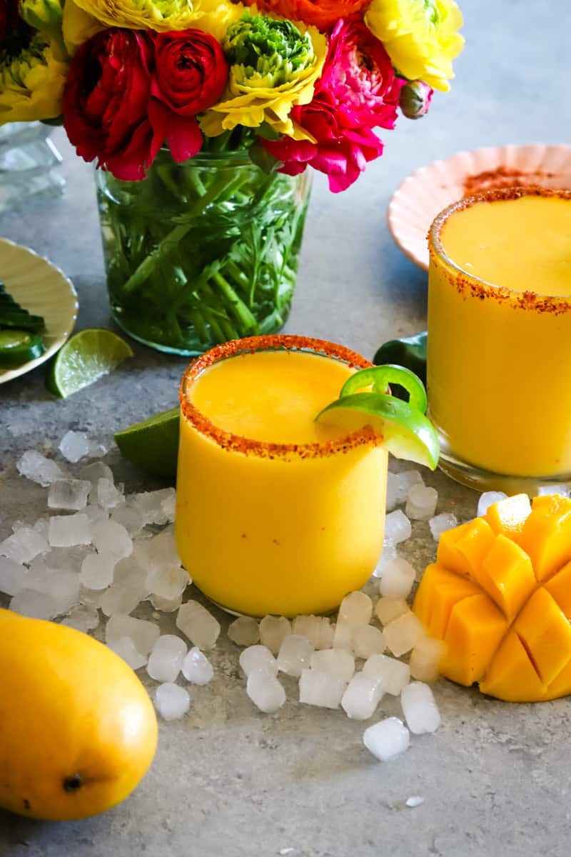 two glasses filled with mango margaritas surrounded by ice cubes and yellow mango. 