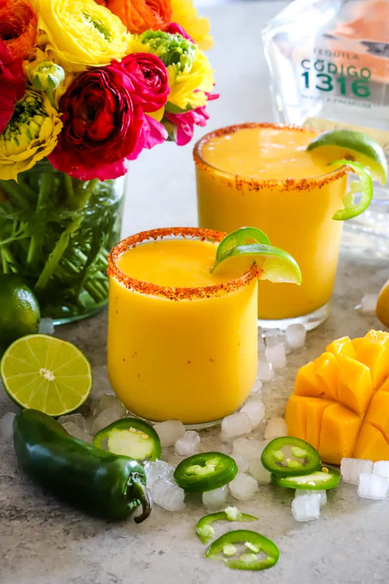 Two glasses of mango margaritas with a vase of flowers in the background and jalapeño slices in the foreground. 