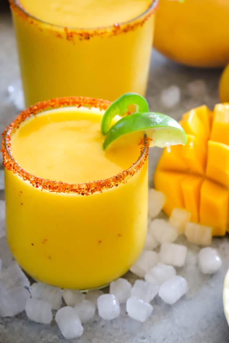 Up close shot of mango margarita with tajin rim and a slice of lime surrounded by ice cubes. 