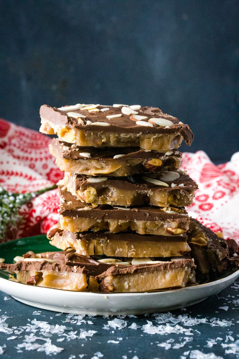 Stacked squares of English butter toffee on green plate with red dish towel behind it. 