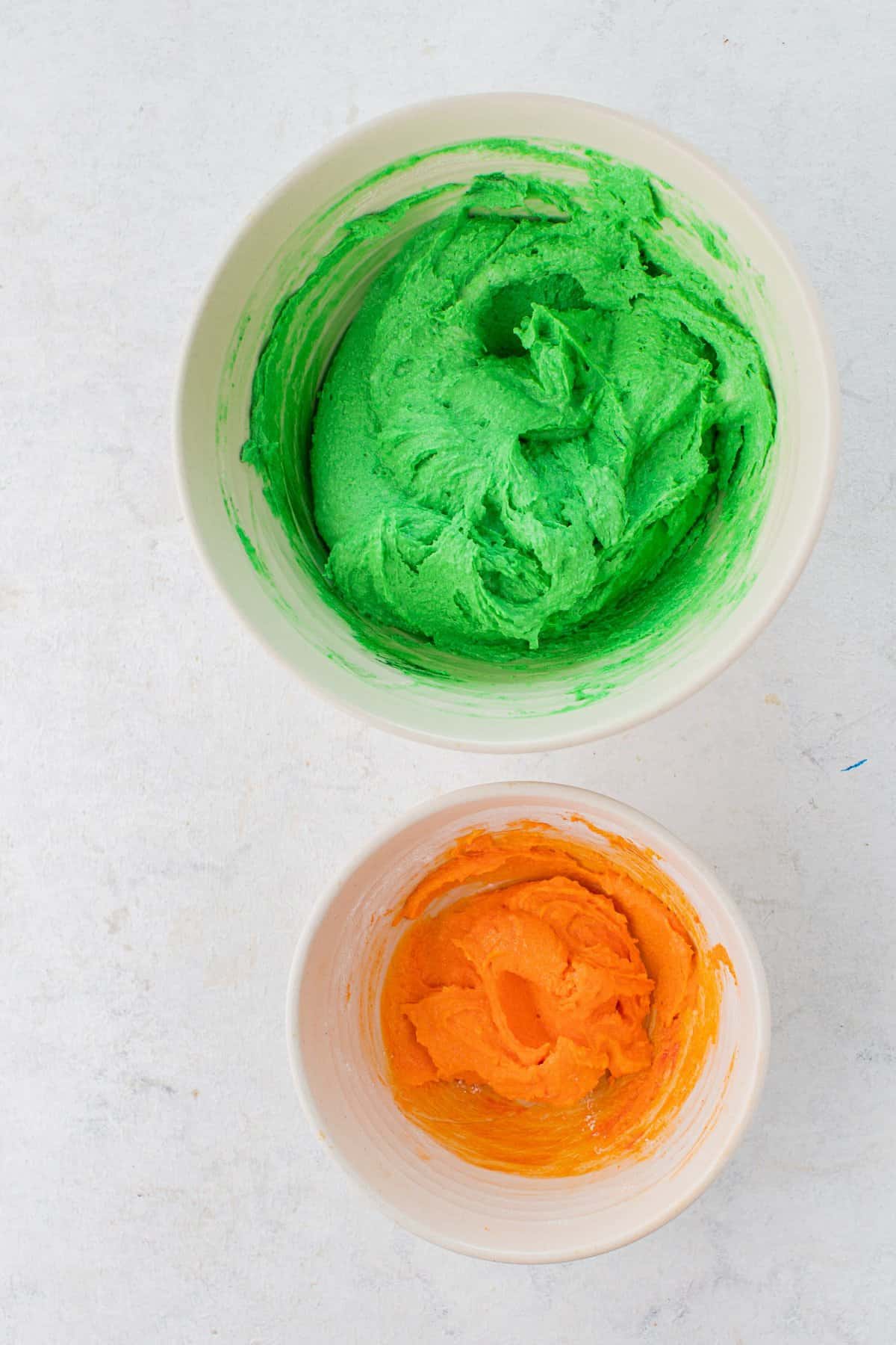 One large white bowl of green frosting next to a small white bowl with orange frosting. 