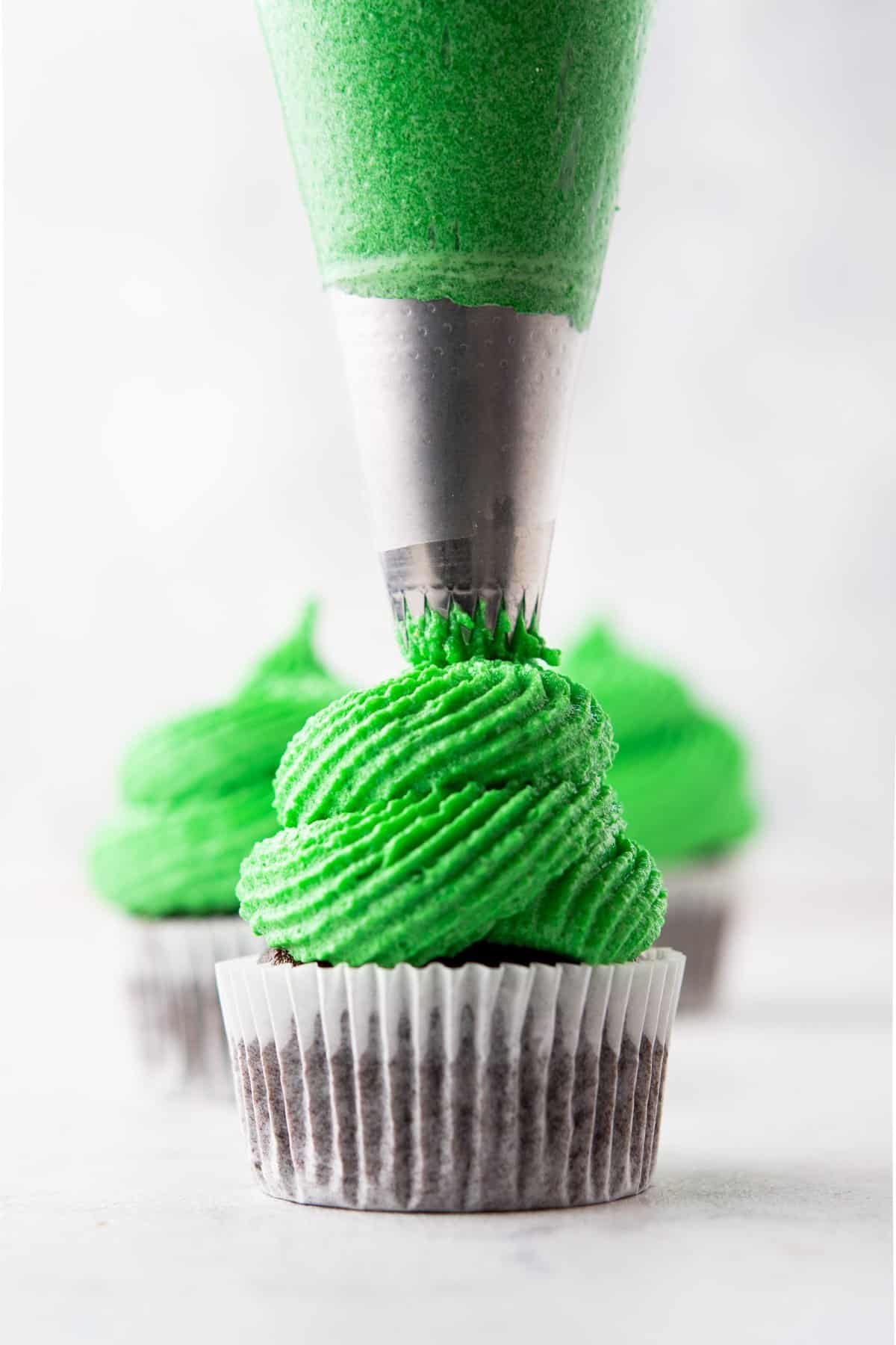 Piping bag frosting the cupcake with green frosting on white surface. 