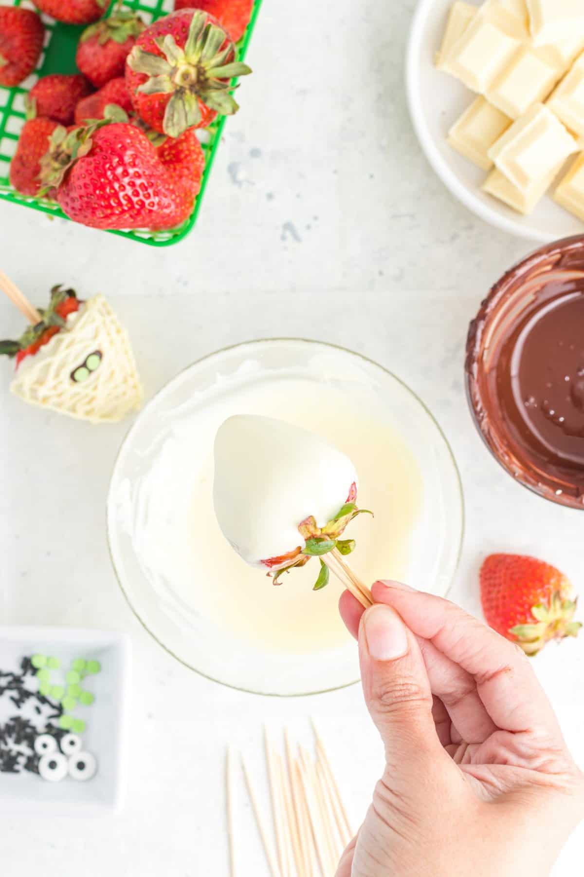 Hand dipping strawberry into a bowl of melted white chocolate. 