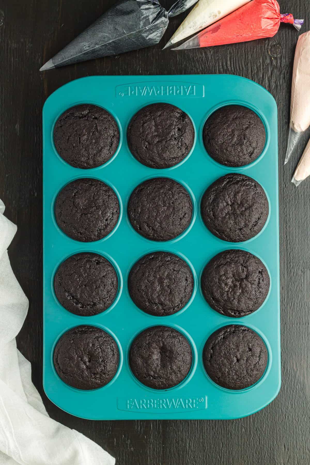 Black cupcakes in a teal muffin pan with bags of frosting on the side.