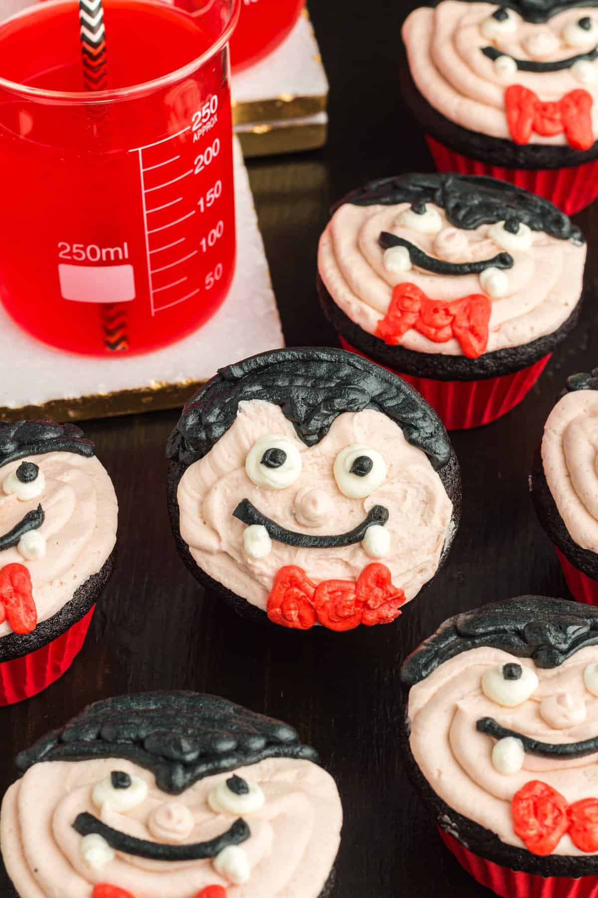 Vampire cupcakes on a black wood background with a jar of red drink to the side. 