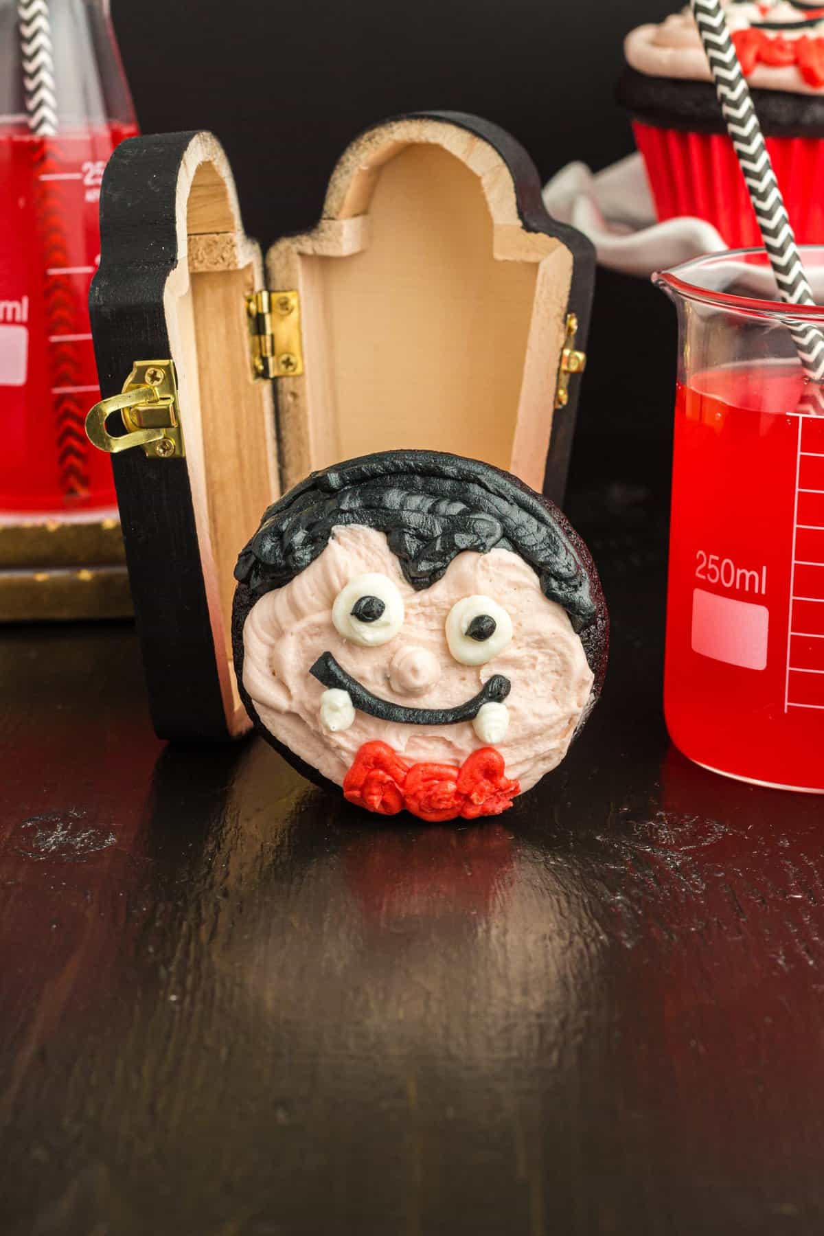 Vampire cupcake laying on its side to show the full vampire face. Wood coffin and red drink in the background.