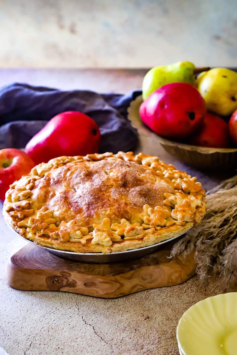 Whole pie on wood plate with bowl of red and green apples and pears in background. 