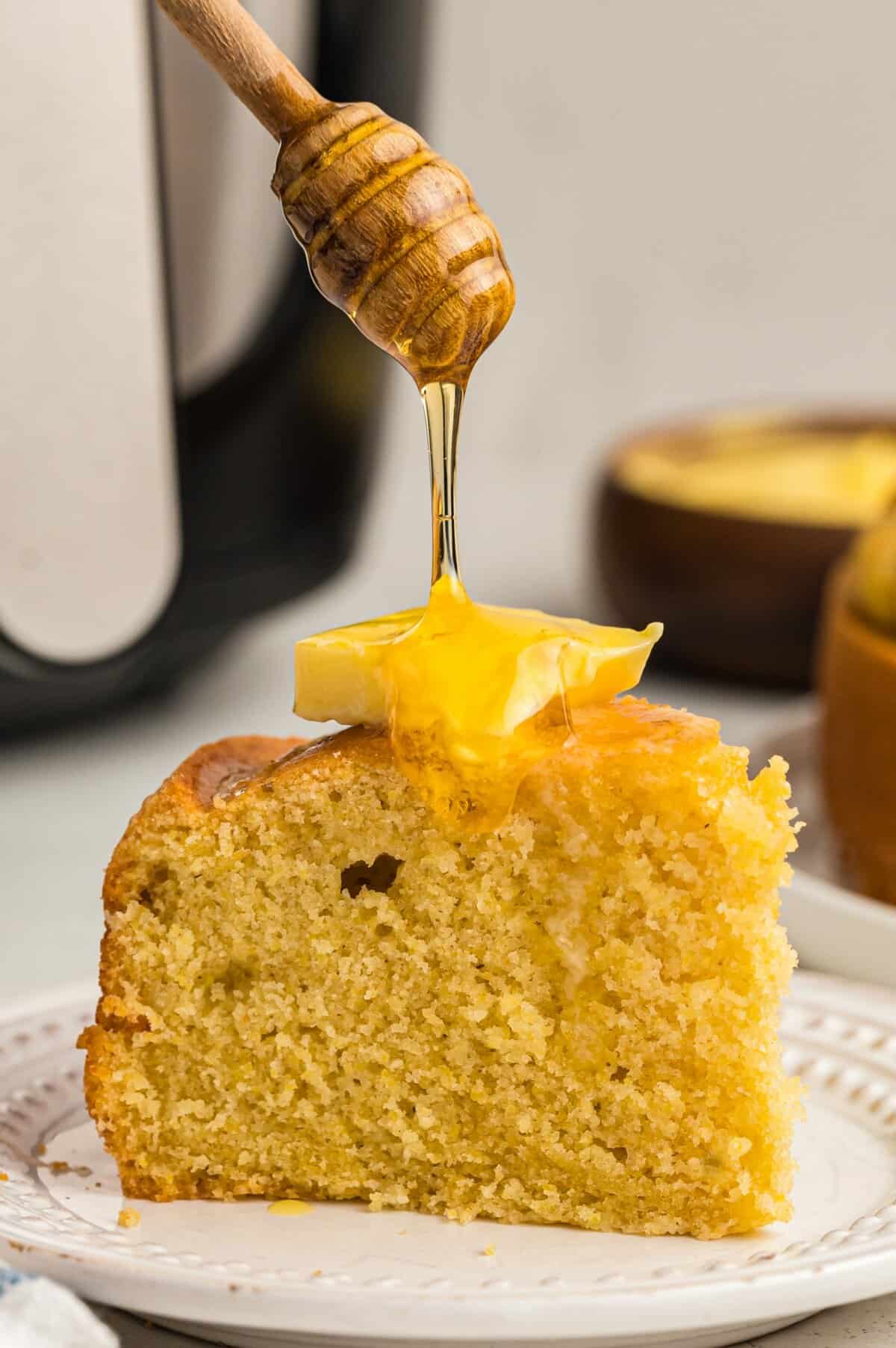 Slice of cornbread on white plate with a pat of butter on top and honey being drizzles. 