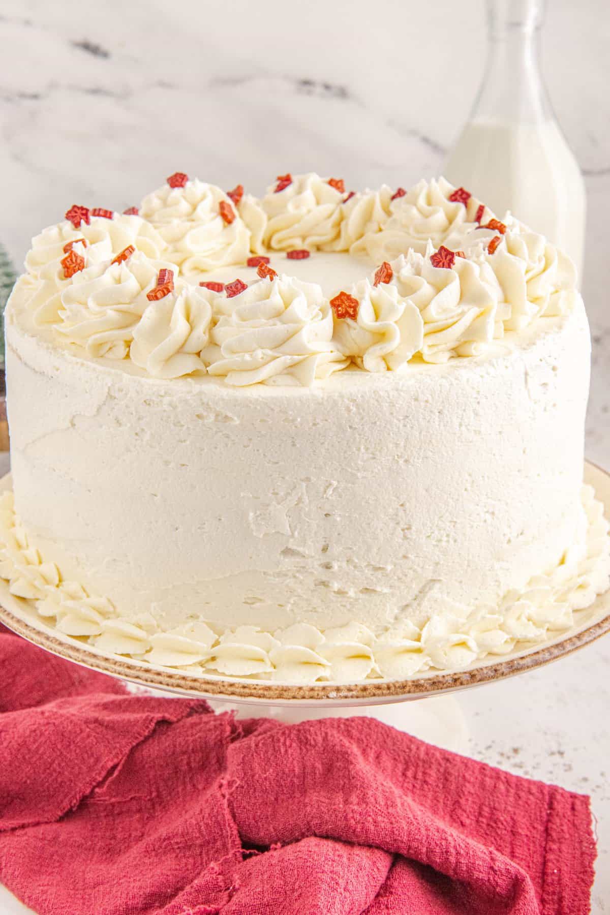 Whole white layer cake on a white cake stand with red kitchen towel in front. 