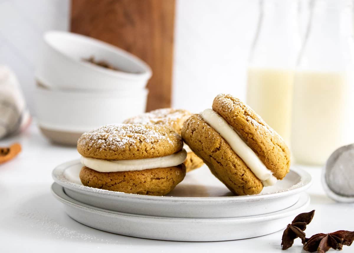 Two cookie sandwiches on a stack of white plates with jugs of milk in the background. 