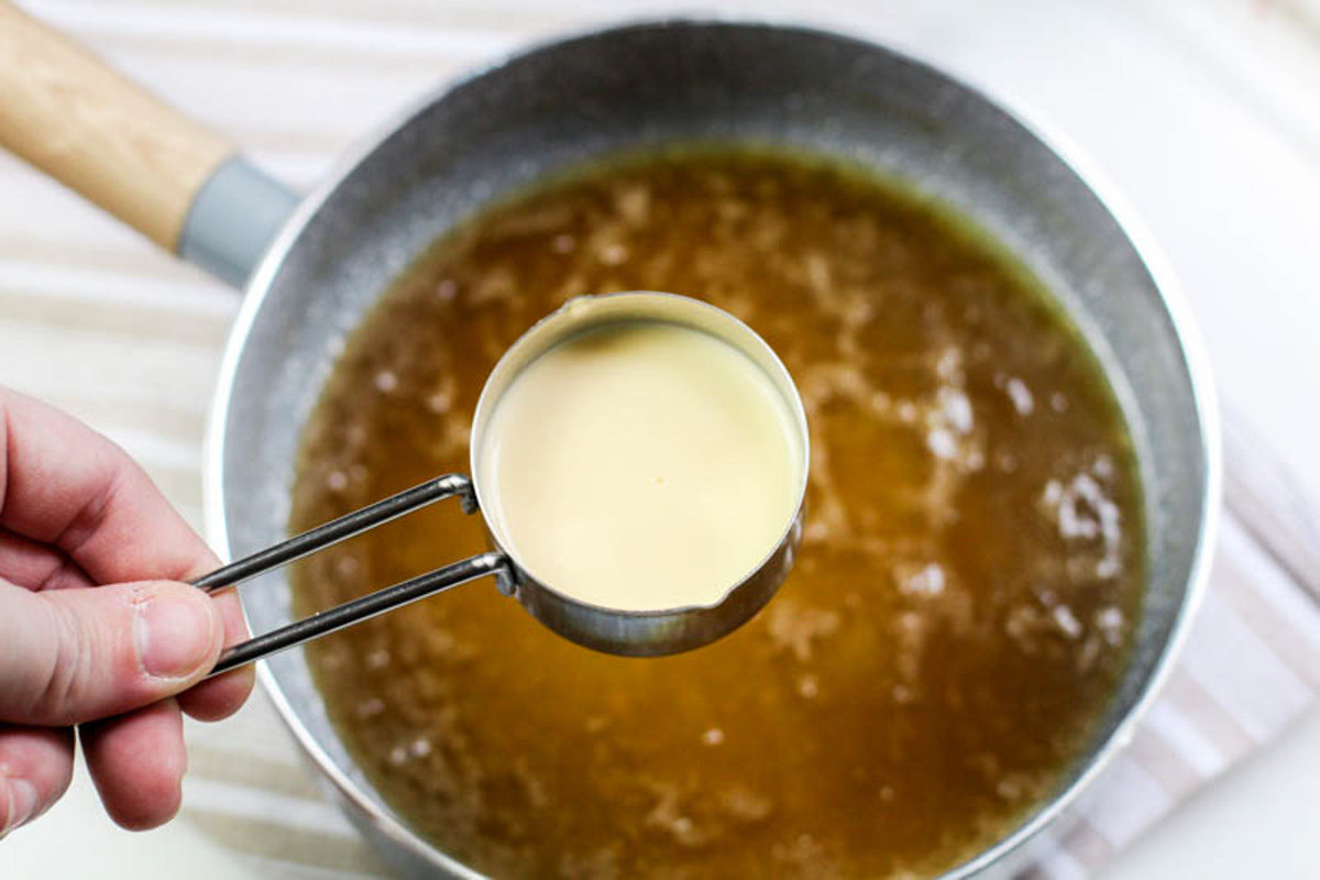 Pouring cream into the caramel mixture in the pot on a white background.