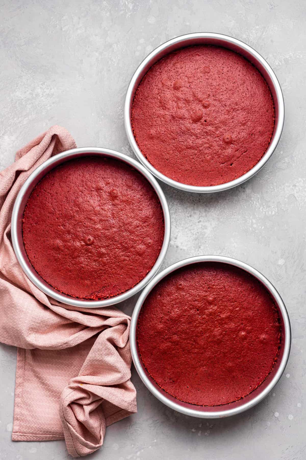 Three round cake pans with the baked red velvet cakes on metal surface. 