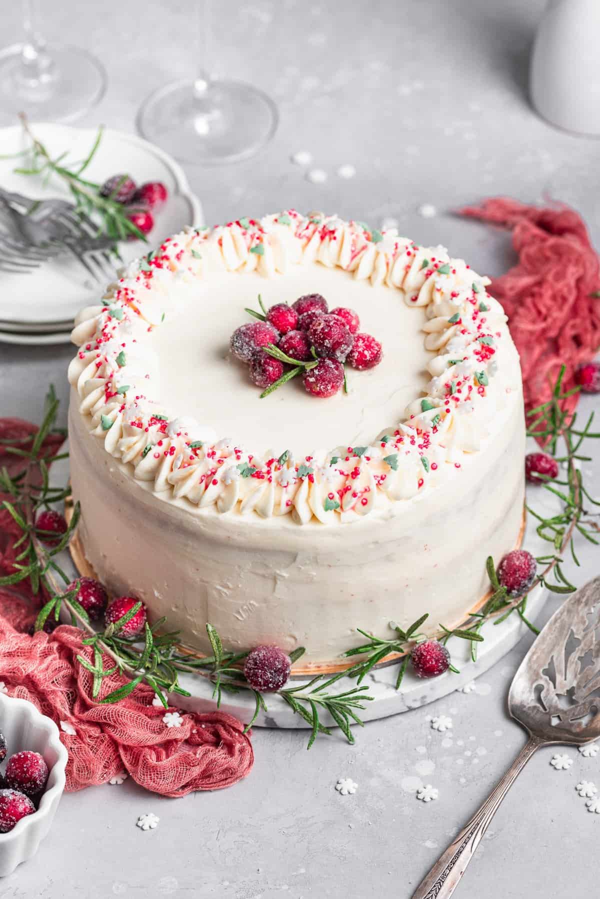 Whole cake with white frosting and sprinkles on white cake plate with rosemary and cranberries. 