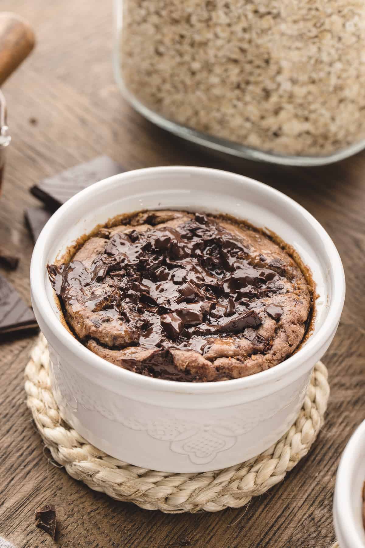 Small white ramekin with chocolate baked oats and jar of oats in background. 