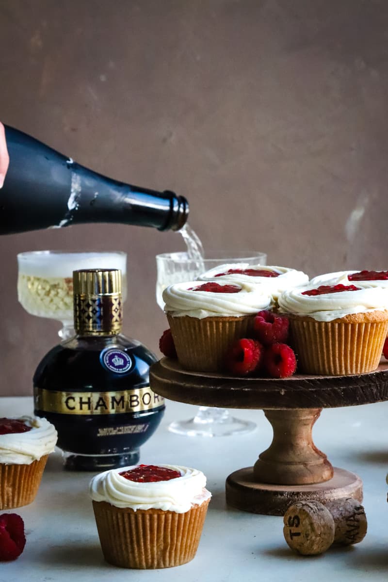 Cupcakes on a wood cutting board with hand pouring a glass of champagne in background. 