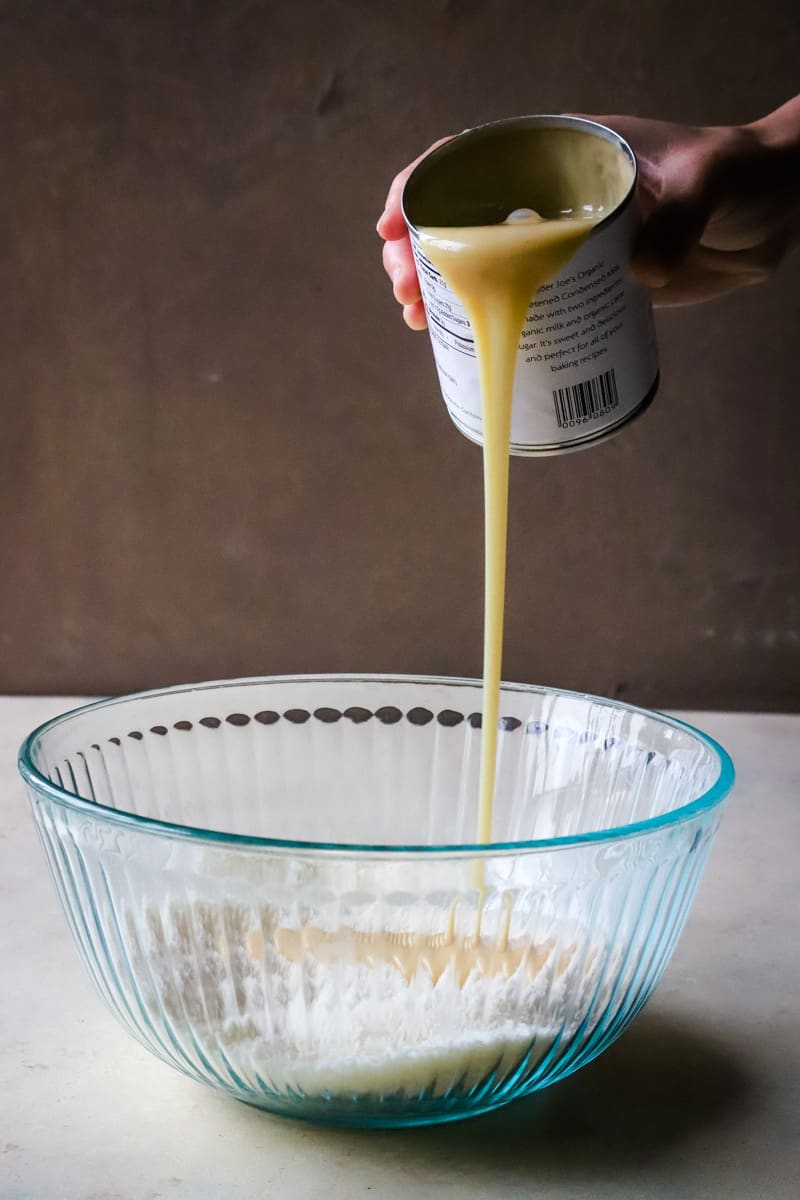 Hand pouring condensed milk into large glass bowl. 