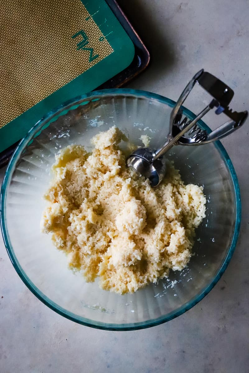 Glass bowl with coconut ball dough and a cookie scoop to roll balls. 