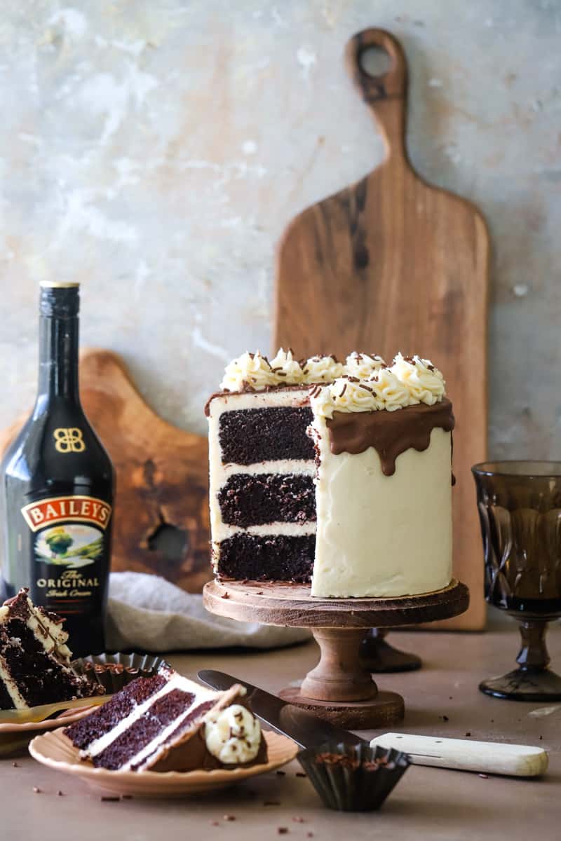 Cake on wood cake stand with baileys bottle and wood cutting boards in background. 