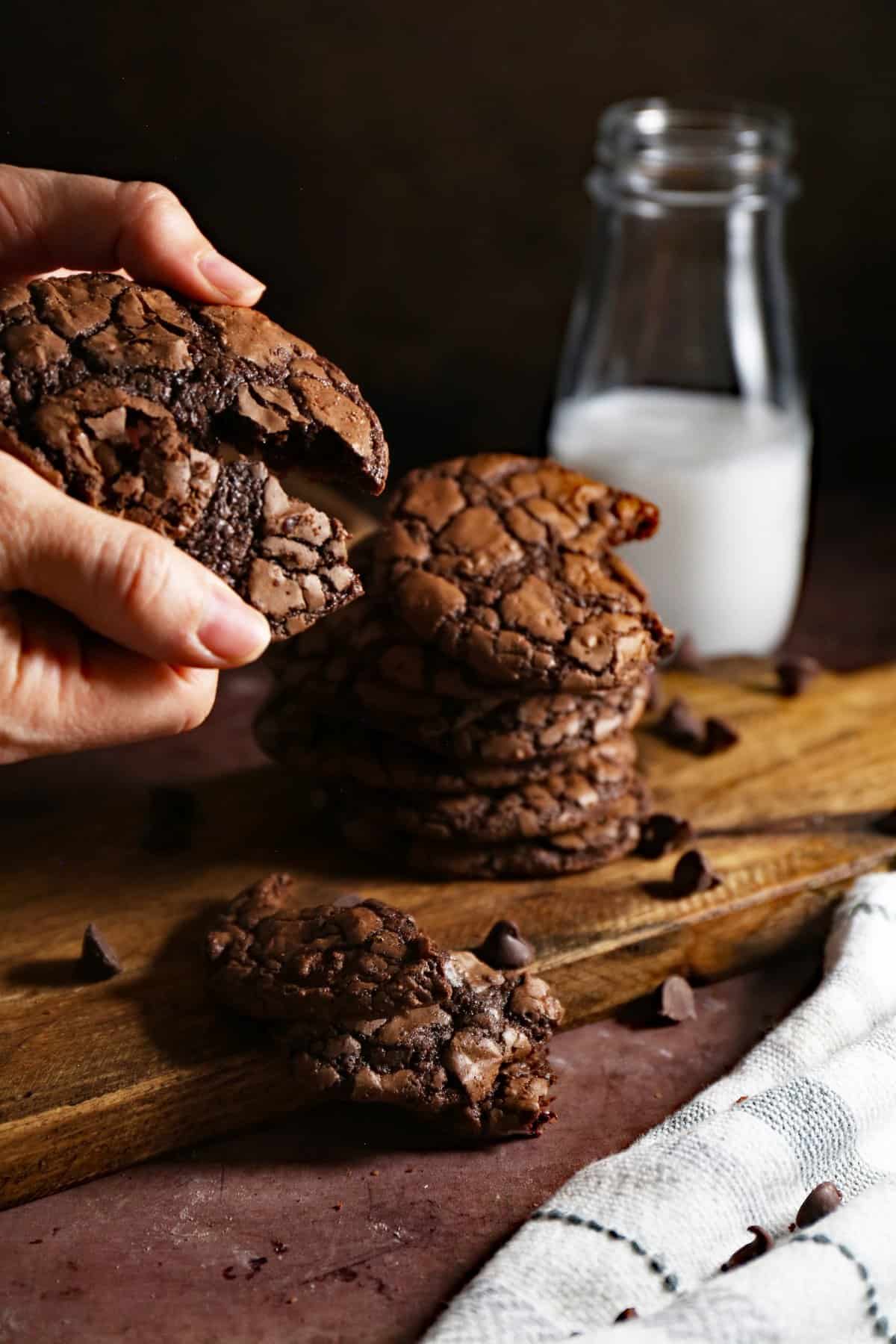 Hand that is breaking apart the chocolate brownie cookies with milk in background. 