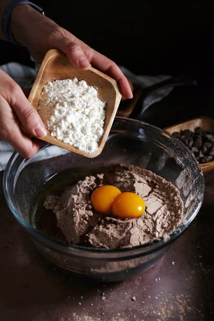 Hand adding additional flour to bowl of eggs and brownie mix