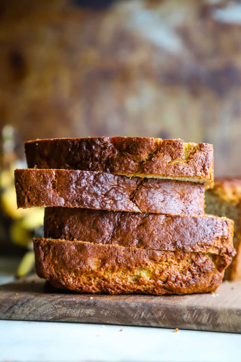 Slices of bread stacked on a wood cutting board with bananas in background. 