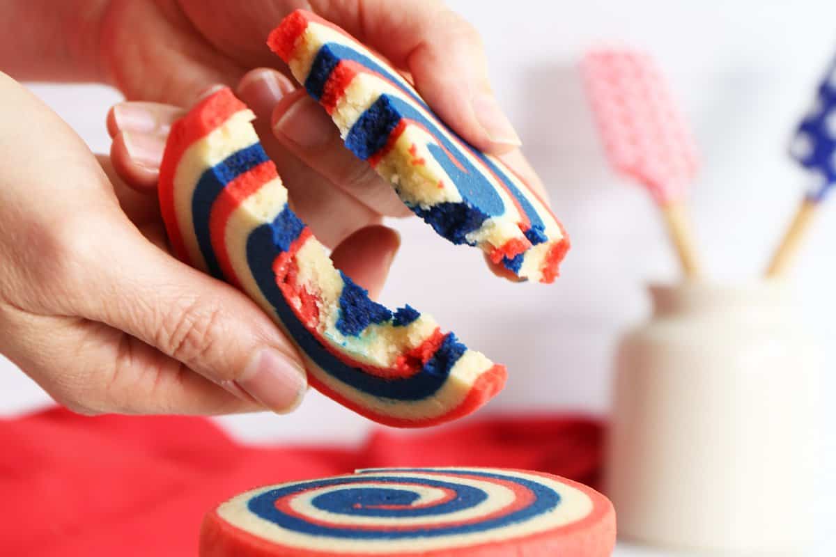 Hand breaking apart a pinwheel cookie with white vase and spatulas in background. 