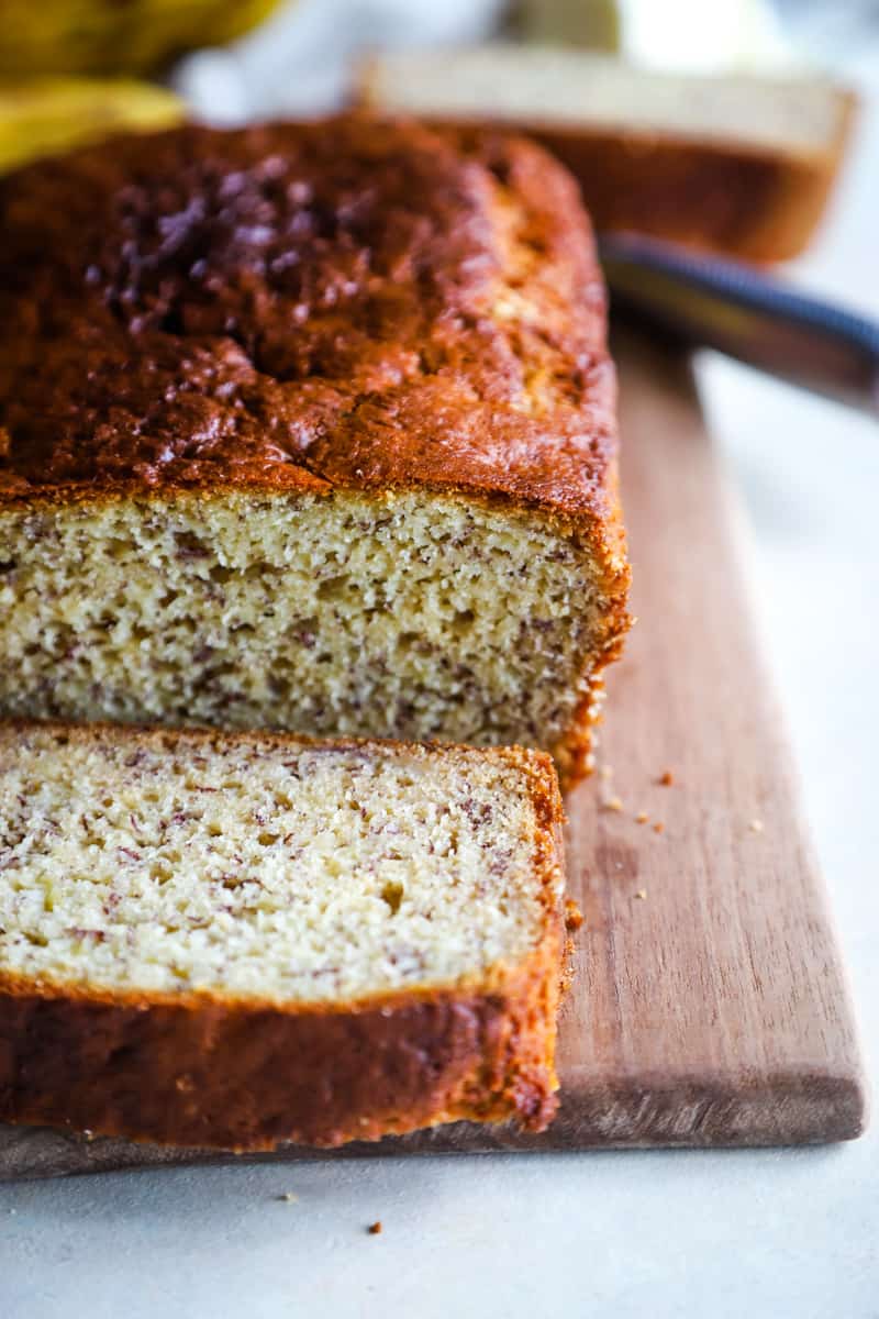 A loaf of banana bread with a slice taken off on a wood cutting board. 