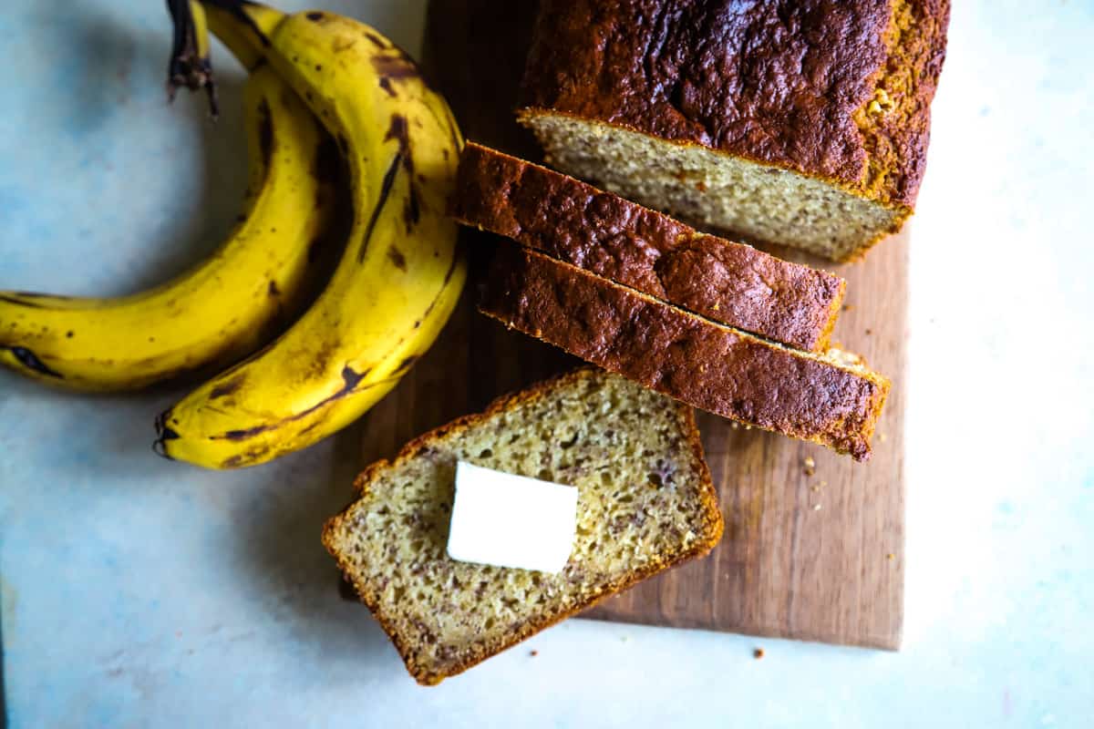 Overhead shot of banana bread slice with pat of butter on wood board. 