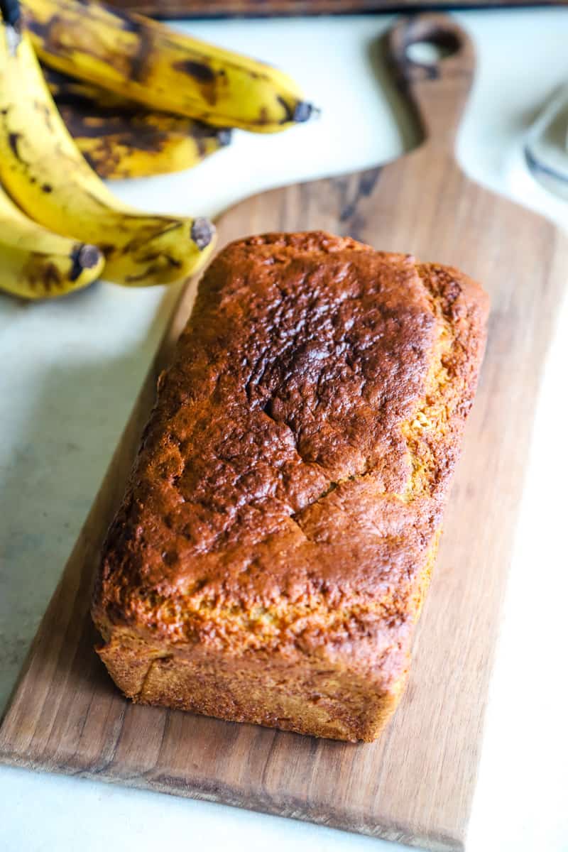 Loaf of banana bread on wood board with bananas in the background. 