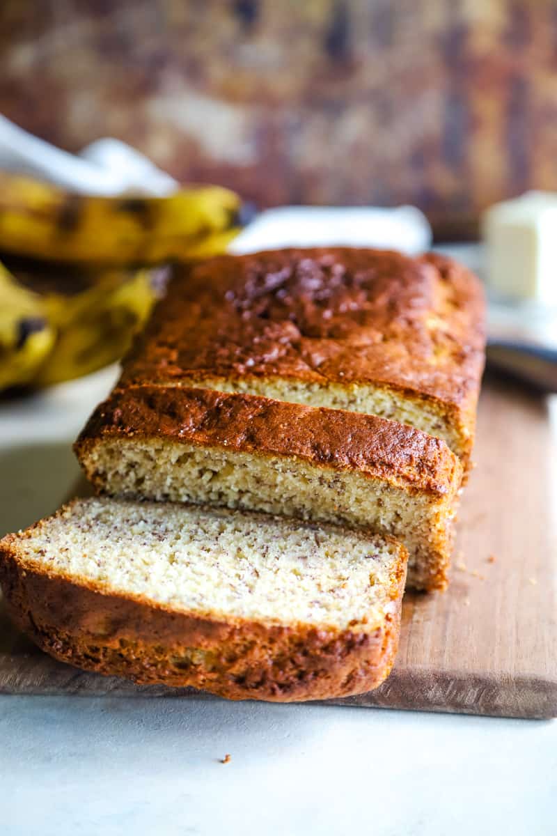 Sliced Banana bread on a wood cutting board with yellow bananas in background. 