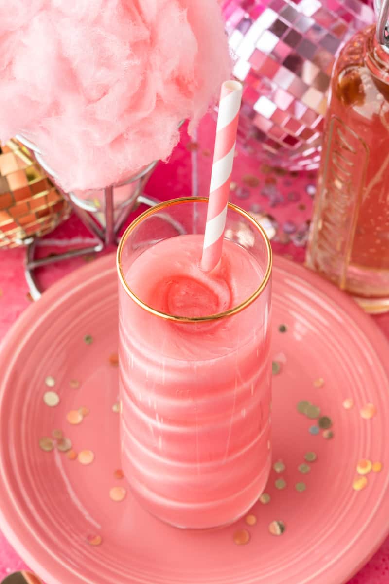 Pink straw in glass filled with pink shimmery cocktail and cotton candy background. 