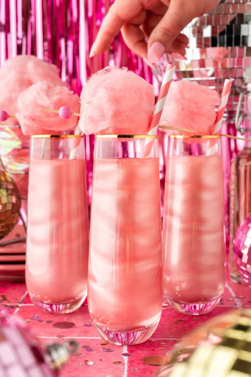 Three pink shimmery cocktails with cotton candy balls on top and a disco ball in background. 