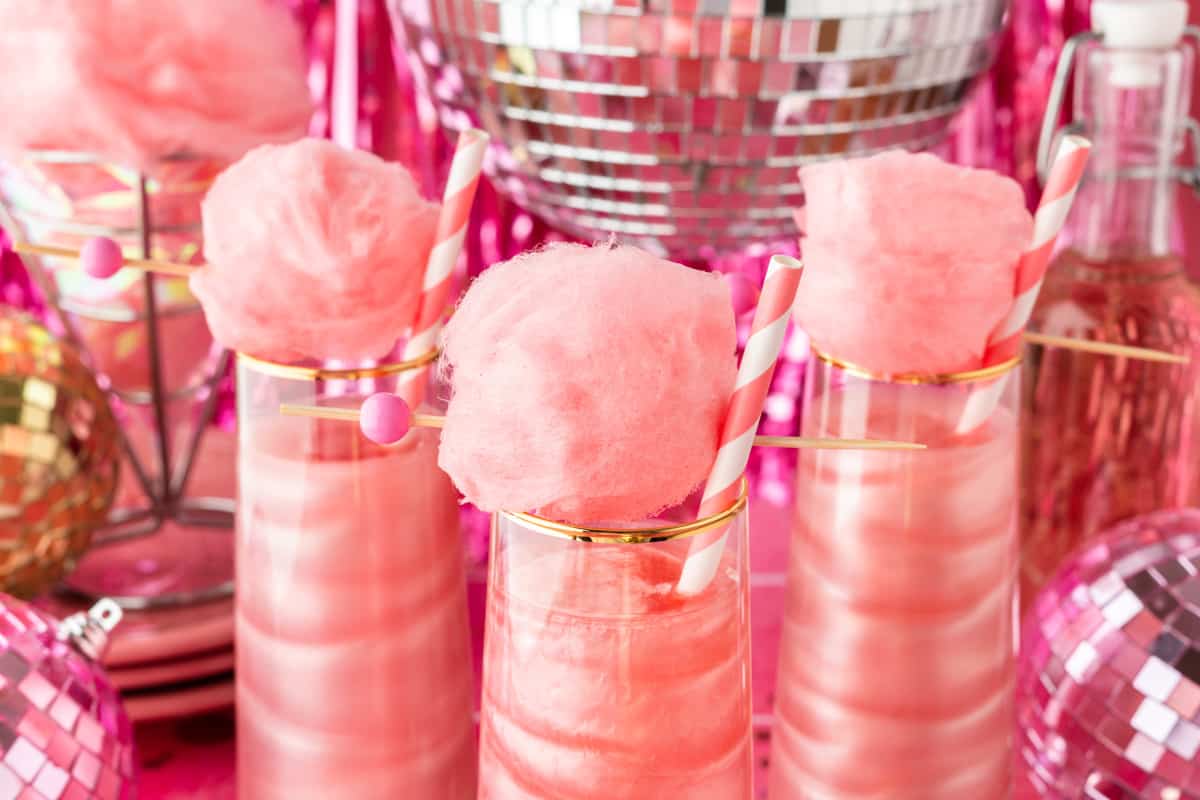 Three cocktails topped with cotton candy and pink straw with disco ball in background. 