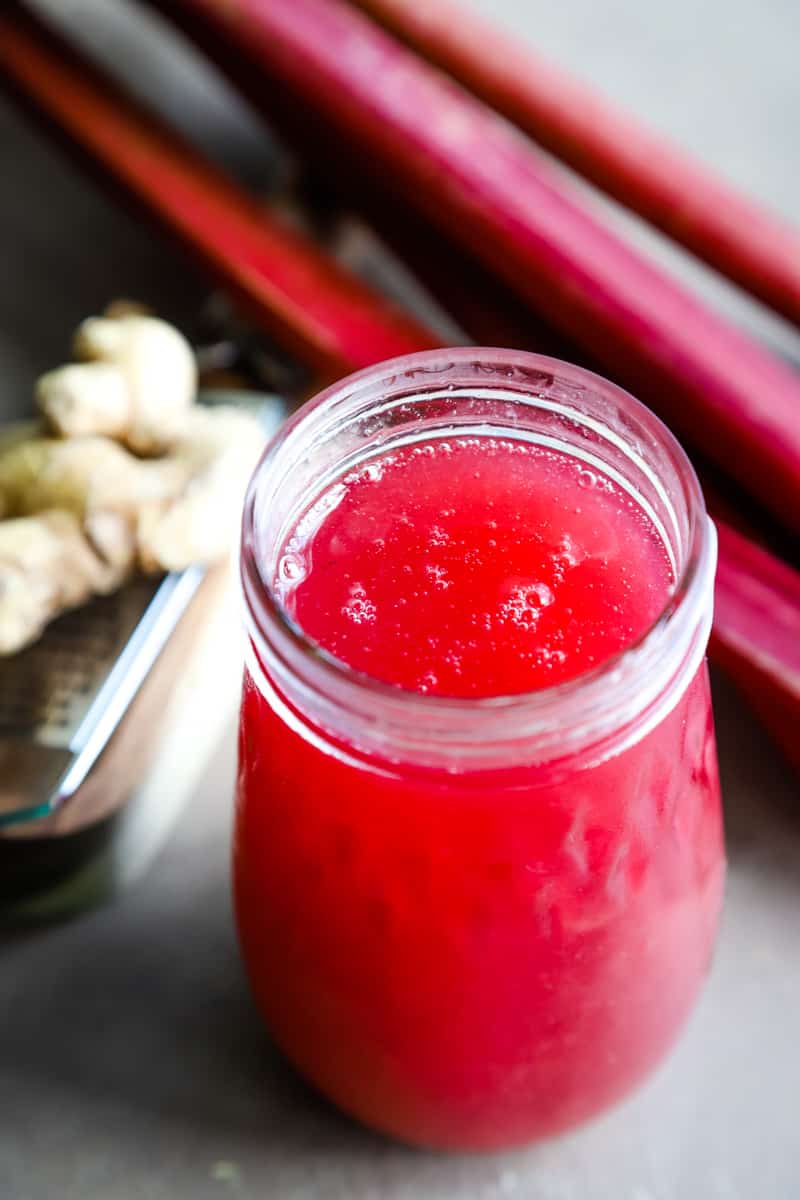 Jar filled with the pink rhubarb syrup and stalks of rhubarb and ginger in background. 
