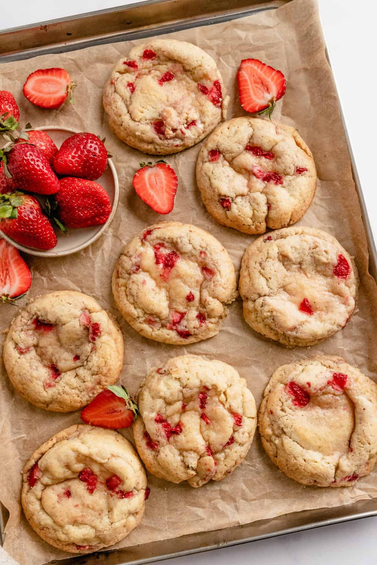 Baked cookies arranged on a parchment lined cookie sheet with bowl of fresh strawberries. 