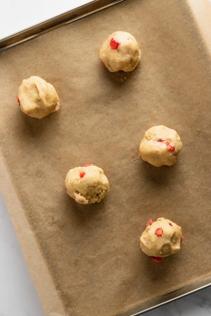 Six Cookie dough balls on a brown parchment lined cookie sheet. 
