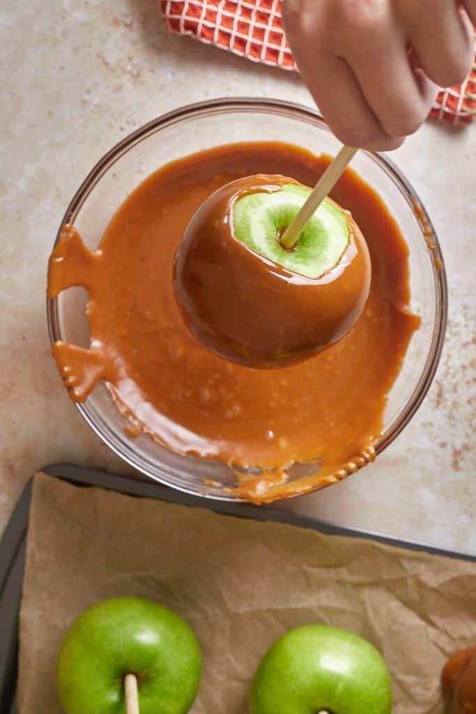 Hand dipping apple on a stick into a glass bowl of melted caramel.  