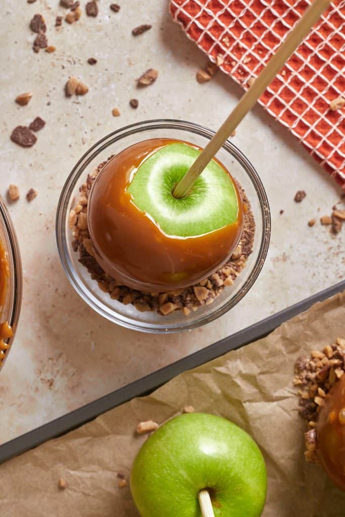 Caramel coated green apple on a stick in a small glass bowl of toffee pieces. 