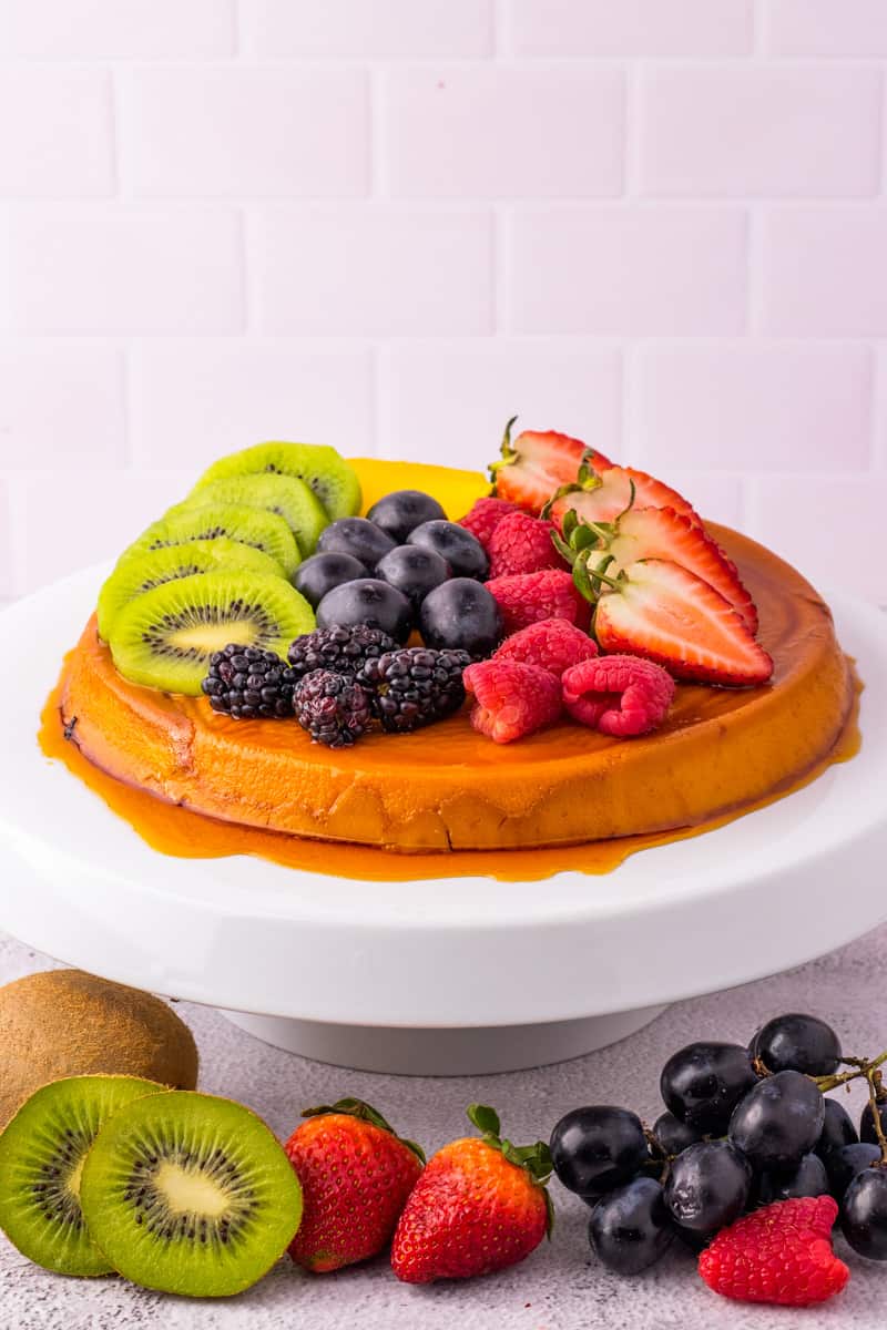 Full flan with kiwis, mango, and berries on top on a white stand. 