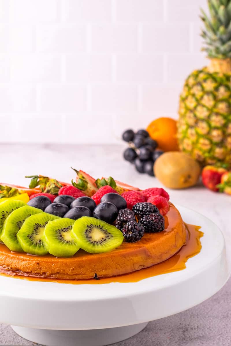 Flan with fruit on a white stand with pineapple and other tropical fruit.