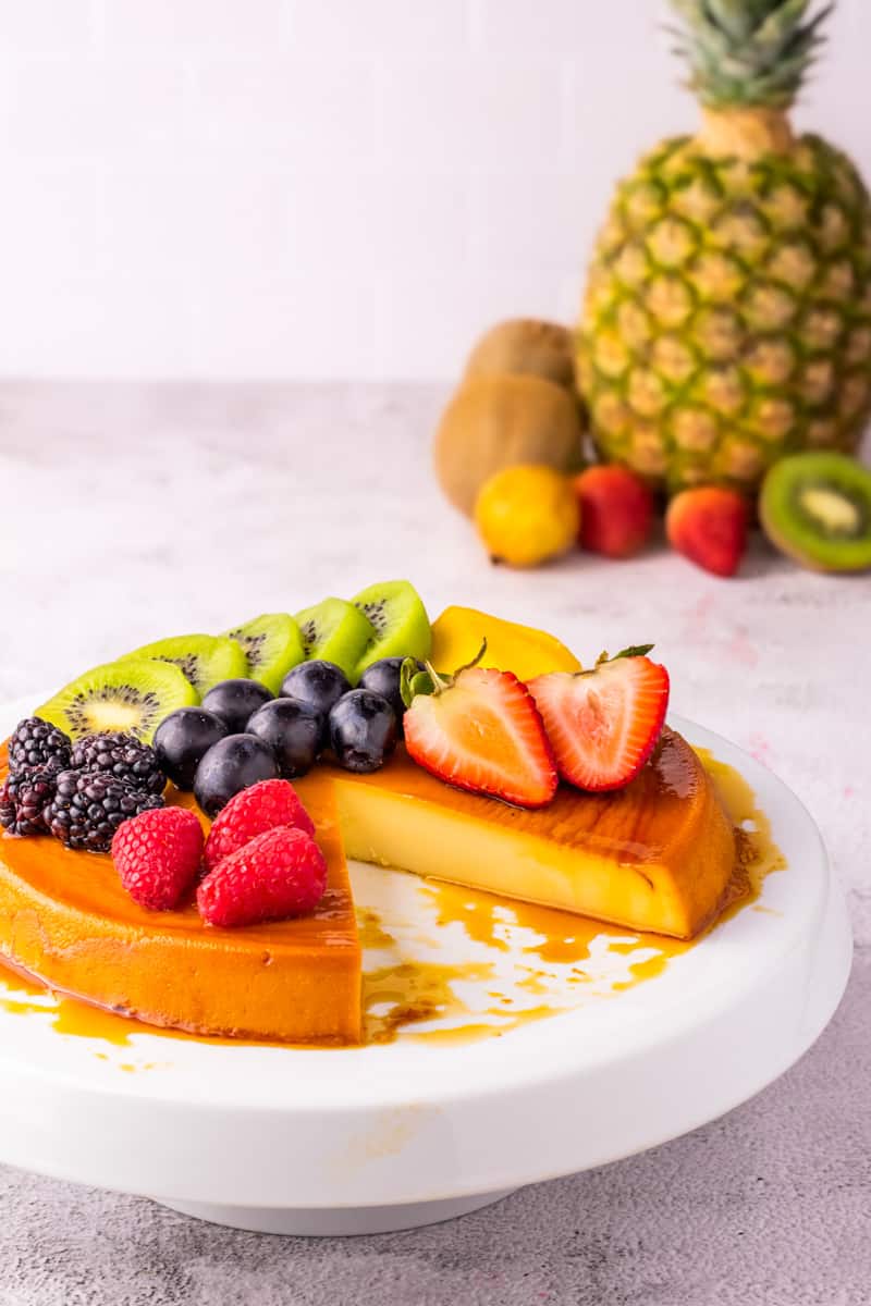 Caramel flan with fruit on top on a white cake stand and white background. 
