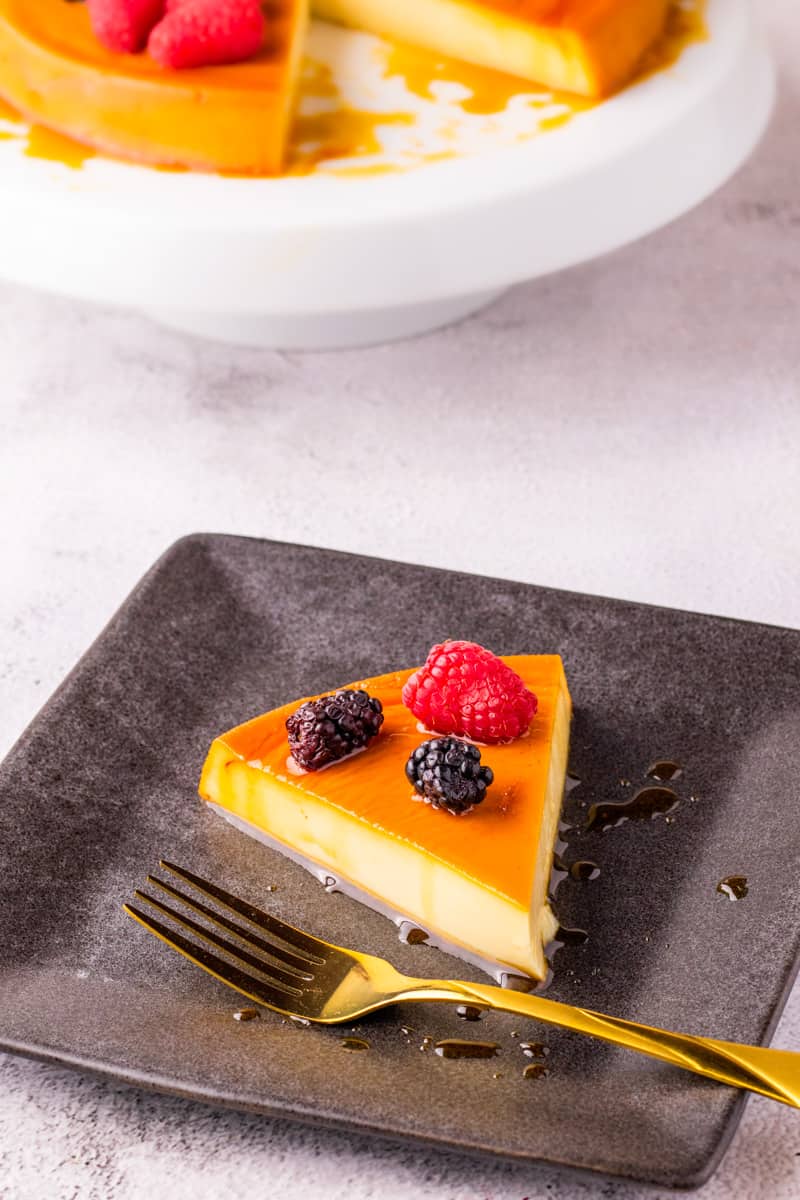 Slice of flan with berries on top on black plate with gold fork. 