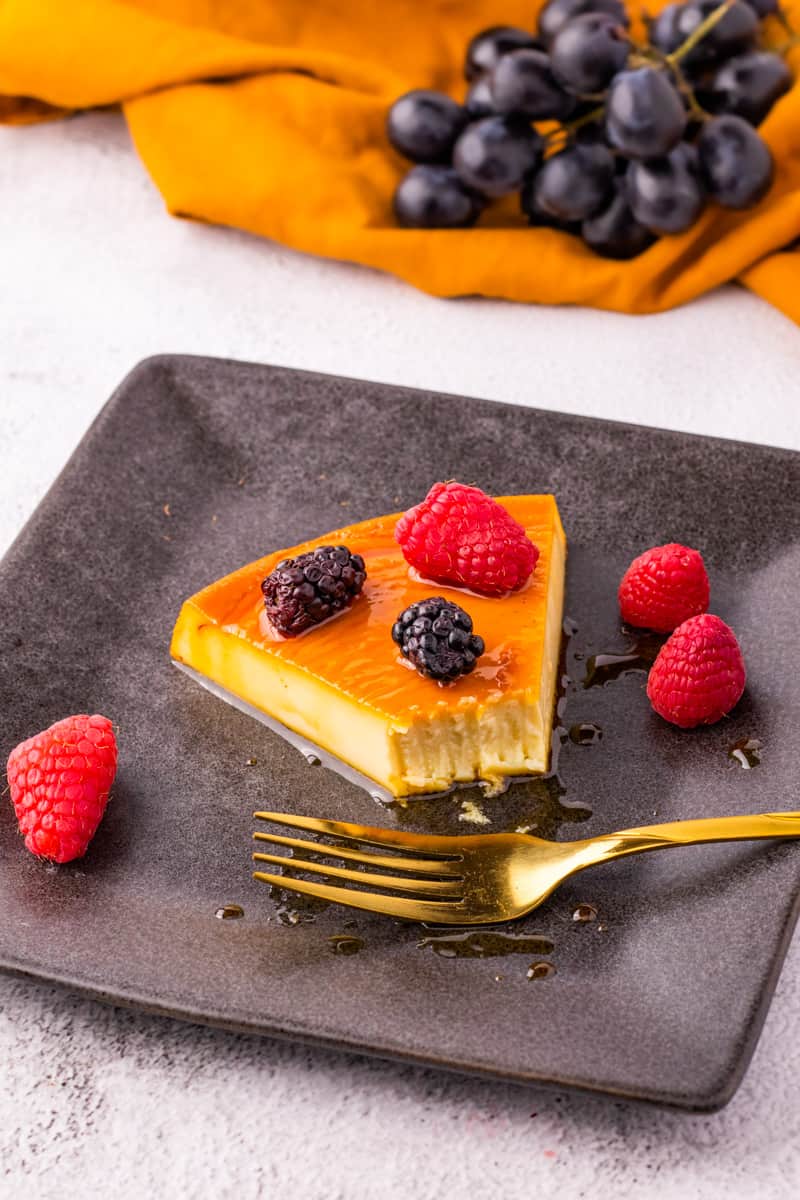 slice of flan cake with fresh berries. on black plate with a gold fork. 