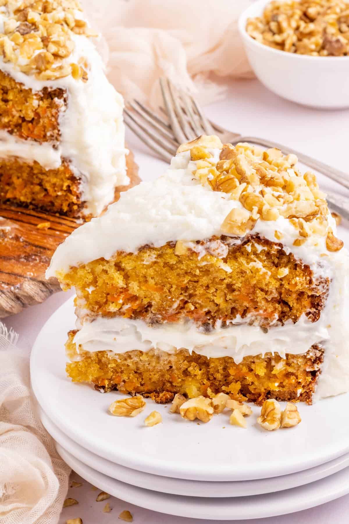 Slice of a layered carrot cake on a stack of white plates with whole cake in background. 